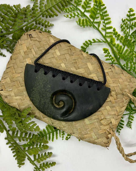 A carved half-circle greenstone pendant with a Māori koru design and bound with a black cord sitting on a woven flax bag amongst green fern leaves.