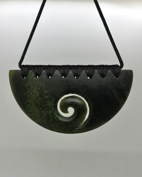 A carved greenstone pendant with a spiral motif, suspended from a black cord against a light grey background.