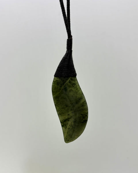 A greenstone pendant carved in the shape of a leaf with natural marbled patterns, wrapped at the top with a woven black cord, shown against a plain grey background.