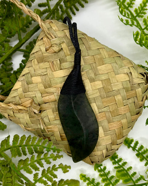 A carved greenstone pendant shaped like a leaf with a black braided cord, displayed on a woven natural fiber pouch surrounded by green fern leaves.