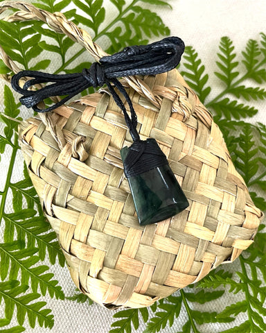 A small, hand carved NZ Greenstone Toki Pendant with a black adjustable woven cord, displayed on a woven flax kete bag. Made from New Zealand Pounamu.