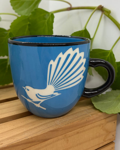 A blue ceramic mug with a black rim and handle, featuring a white silhouette of a Fantail bird, displayed on a wooden slatted surface with green leaves in the background.
