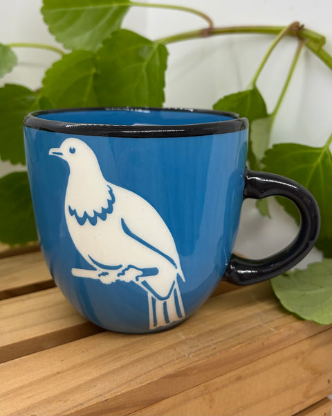 A blue ceramic mug with a black handle and rim, featuring a white kererū bird design, set on a wooden slatted surface with green leaves in the background.