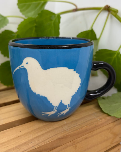 A blue ceramic mug with a black handle and rim, featuring a white kiwi bird illustration on the side, set on a wooden slatted surface with green leaves in the background.