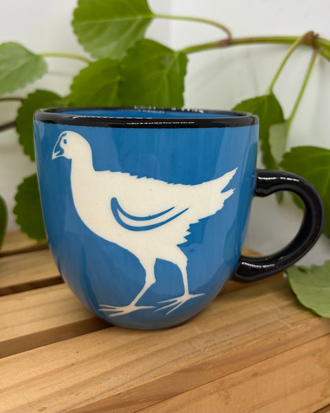 A blue ceramic mug with a black handle and rim, featuring a white pūkeko bird silhouette design, set on a wooden surface with green leaves in the background.