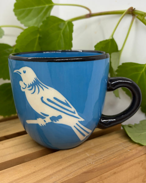 A blue ceramic mug with a black handle and rim, featuring a white tūī bird silhouette printed on the side, resting on a wooden surface with green leaves in the background.