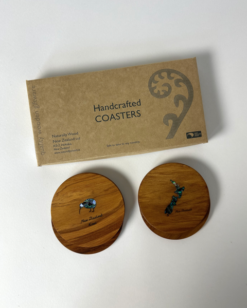 A set of four round, wooden coasters with pāua shell inlay. One coaster features a kiwi bird design and the other with a New Zealand map design, placed on a surface with a cardboard gift box in the background that reads 'Handcrafted Coasters'.