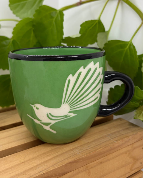 A green ceramic mug with a black rim and handle, featuring a white silhouette of a Fantail bird, set on a wooden surface with green leaves in the background.