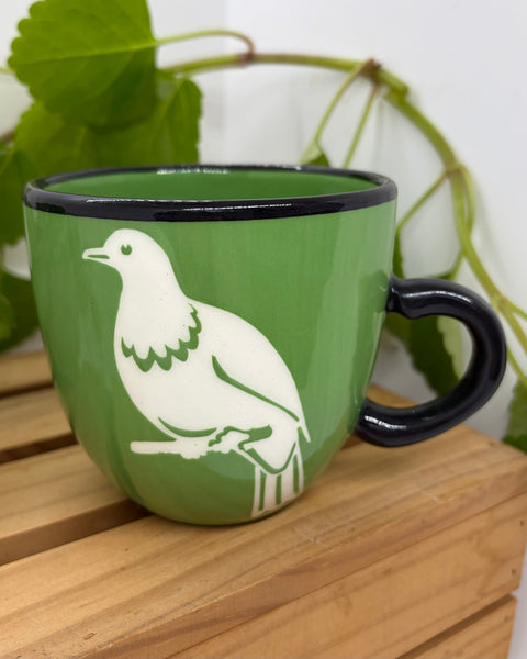 A green ceramic mug with a black rim and handle, featuring a white silhouette of a kererū bird perched on a branch, displayed on a wooden slatted surface with green leaves in the background.