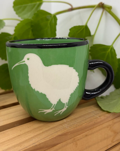 A green ceramic mug with a black rim and handle, featuring a white kiwi bird illustration, displayed on a wooden slatted surface with green leaves in the background.