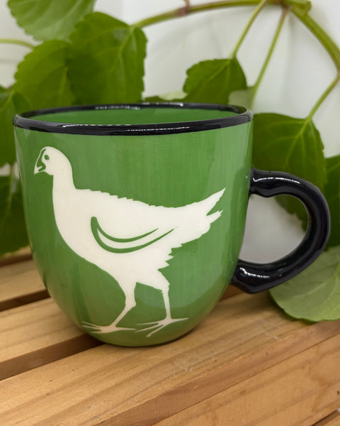A green ceramic mug with a black rim and handle, featuring a white pūkeko bird silhouette design, displayed on a wooden surface with green leaves in the background.