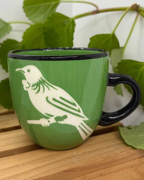 A green ceramic mug with a black handle and rim, featuring a white tūī bird illustration on the side, set on a wooden surface with green leaves in the background.