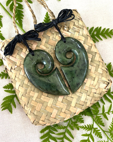 A two piece set of New Zealand Greenstone koru pendants that, when aligned, form two halves of a heart. With black woven cords and displayed on a flax kete bag.