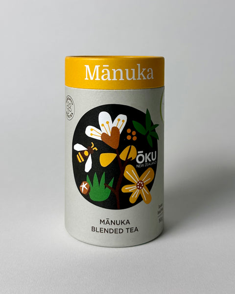 A cylindrical container of OKU Manuka blended loose leaf tea with illustrations of manuka flowers and leaves, and text indicating the product name and type.