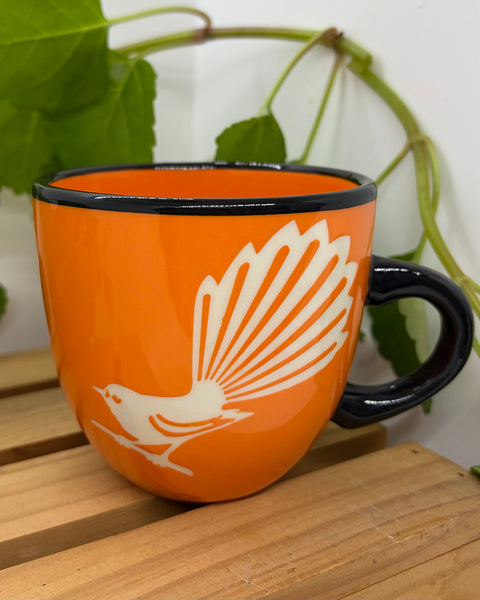 An orange ceramic mug with a black rim and handle, featuring a white graphic of a Fantail bird perched on a branch, displayed on a wooden surface with green plant leaves in the background.