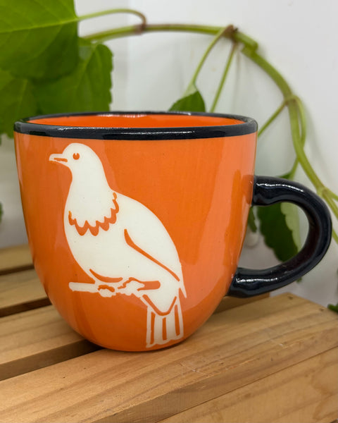An orange ceramic mug with a black handle and rim, featuring a white kererū bird silhouette printed on the side, set on a wooden surface with green leaves in the background