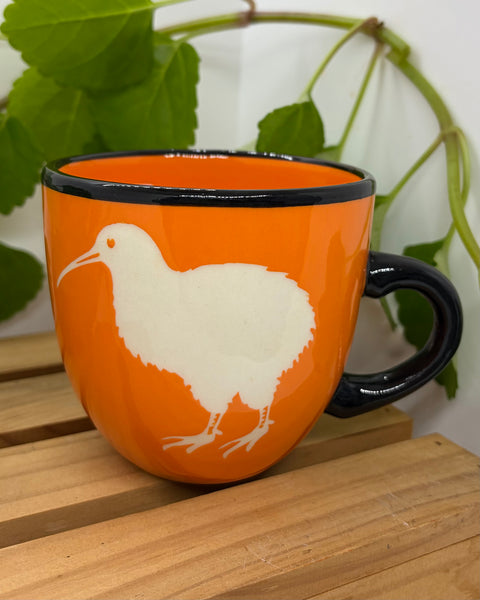 An orange ceramic mug with a black handle and rim, featuring a white kiwi bird illustration, displayed on a wooden slatted surface with green leaves in the background.