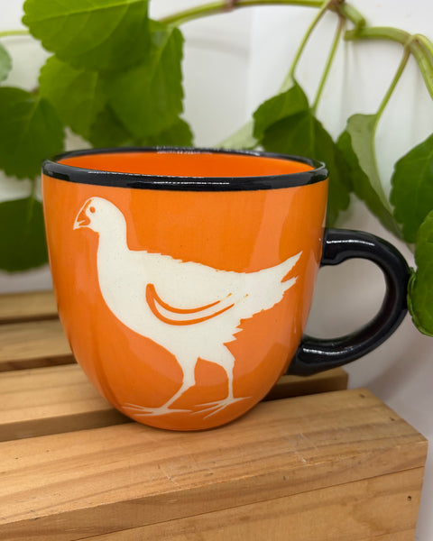 An orange ceramic mug with a black rim and handle, featuring a white pūkeko silhouette printed on the side, displayed on a wooden surface with green leaves in the background.