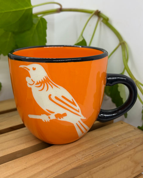 An orange ceramic mug with a black rim and handle, featuring a white tūī bird illustration on the side, resting on a wooden slatted surface with green leaves in the background.