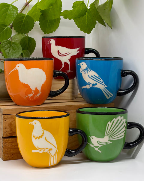 Five brightly coloured ceramic mugs with black handles and rims, each decorated with a different bird illustration in white. The mugs are arranged on wooden blocks against a white wall with green leaves hanging in the background.