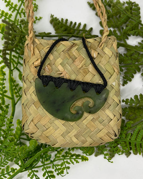 A carved greenstone pendant with two spiral cutouts, bound with a woven black cord, on top of a woven flax bag amongst green fern fronds.