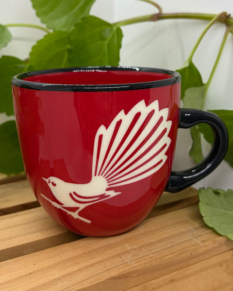 A red ceramic mug with a black handle and rim, featuring a white Fantail bird design on the front, set on a wooden surface with green leaves in the background.