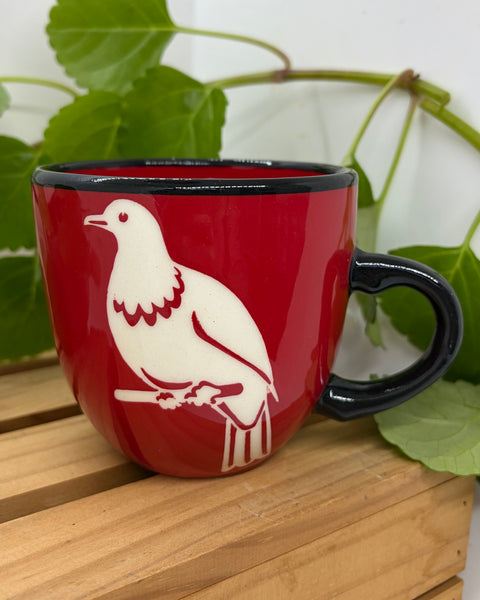 A red ceramic mug with a black handle and rim, featuring a white kererū perched on a branch printed on the side. The mug is placed on a light wooden slatted surface with green leaves in the background.