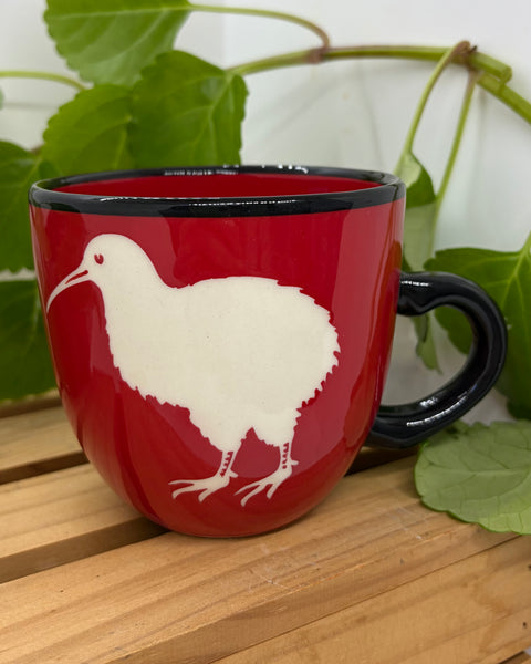 A red ceramic mug with a black handle and rim, featuring a white kiwi bird illustration on the side, displayed on a wooden surface with green leaves in the background.