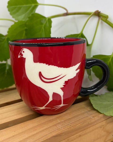 A red ceramic mug with a black handle and rim, featuring a white pūkeko bird silhouette design on the side, set on a wooden surface with green leaves in the background.