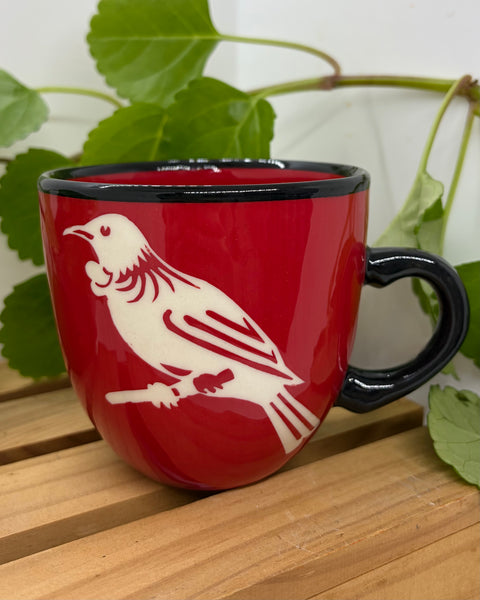 A red ceramic mug with a black handle and rim, featuring a white-coloured tui bird perched on a branch on the front. The mug is placed on a light wooden surface with green leaves in the background.