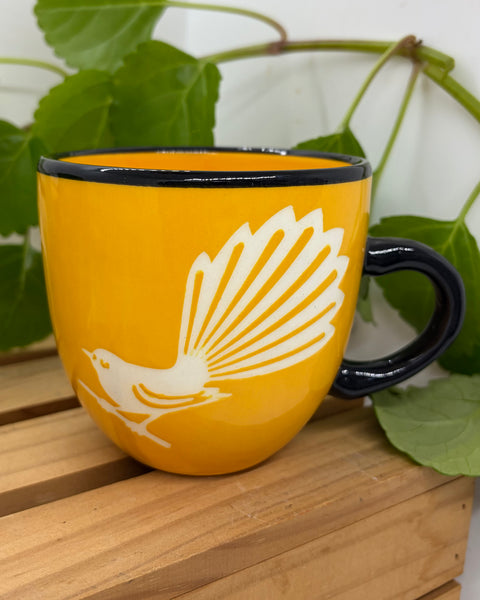 A yellow ceramic mug with a black rim and handle, featuring a white silhouette of a Fantail bird perched on a branch, displayed on a wooden slatted surface with green leaves in the background.