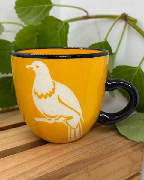 A yellow ceramic mug with a black rim and handle, featuring a white silhouette of a kererū bird perched on a branch, set on a wooden surface with green leaves in the background.