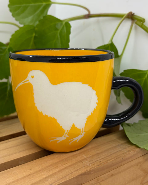 A yellow ceramic mug with a black handle and rim, featuring a white kiwi bird silhouette, set on a wooden surface with green leaves in the background.