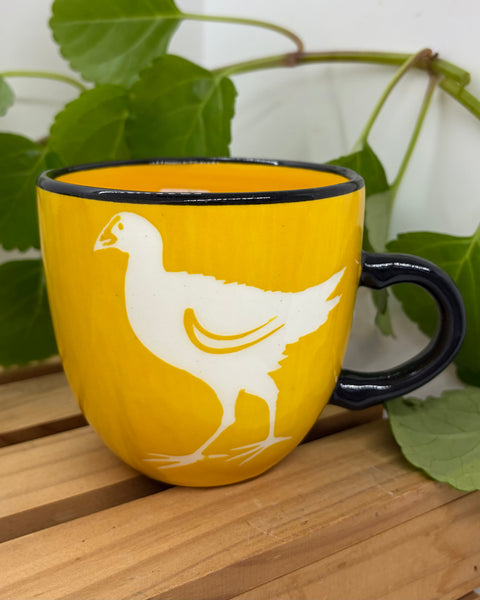 A yellow ceramic mug with a black handle and rim, featuring a white silhouette of a pūkeko bird on the side. The mug is placed on a wooden slatted surface with green leaves in the background.