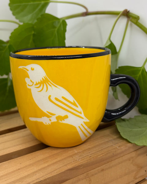 A yellow ceramic mug with a black rim and handle, featuring a white silhouette of a tūī bird perched on a branch, set on a wooden surface with green leaves in the background.